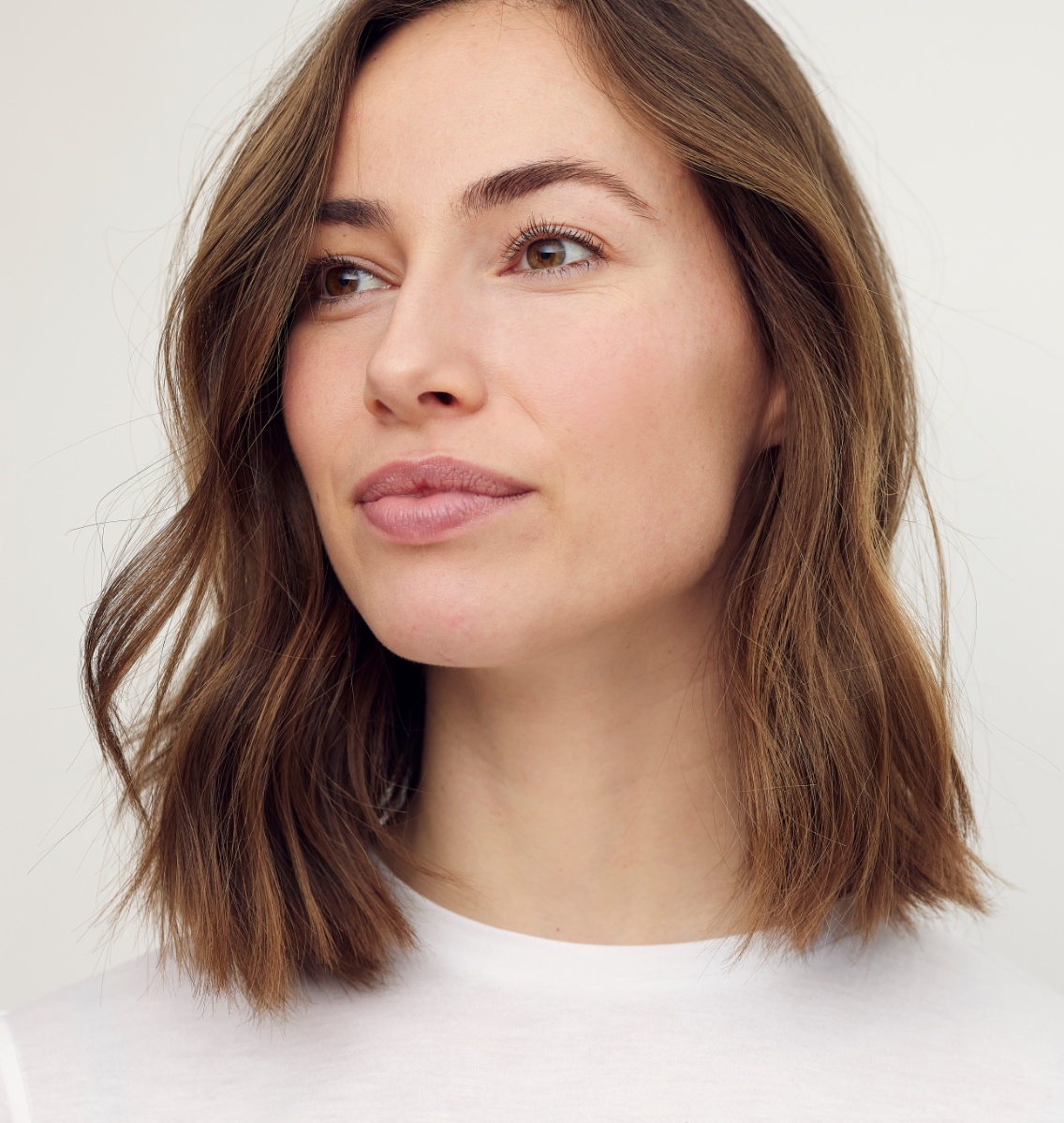 Woman with shoulder-length brown hair and hazel eyes wearing a white top, gazing thoughtfully to the side