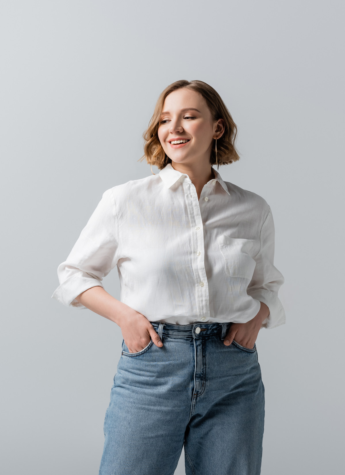 Smiling woman with short wavy hair wearing a white button-up shirt and blue jeans, standing against a light gray background with hands in her pockets.