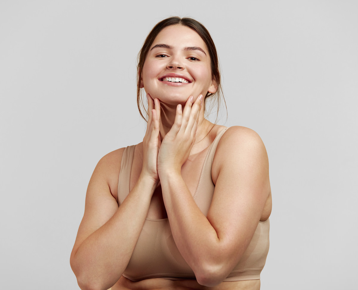 Smiling woman in a beige tank top gently touching her face with both hands against a plain light gray background.