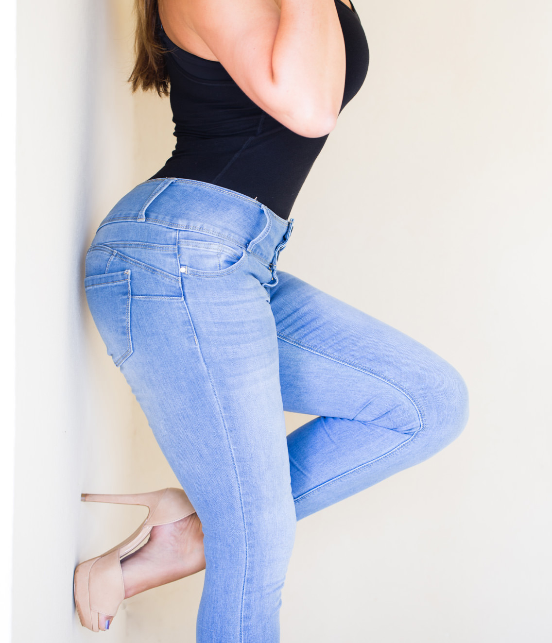 Woman wearing light blue skinny jeans, a black sleeveless top, and nude heels posing sideways against a wall.